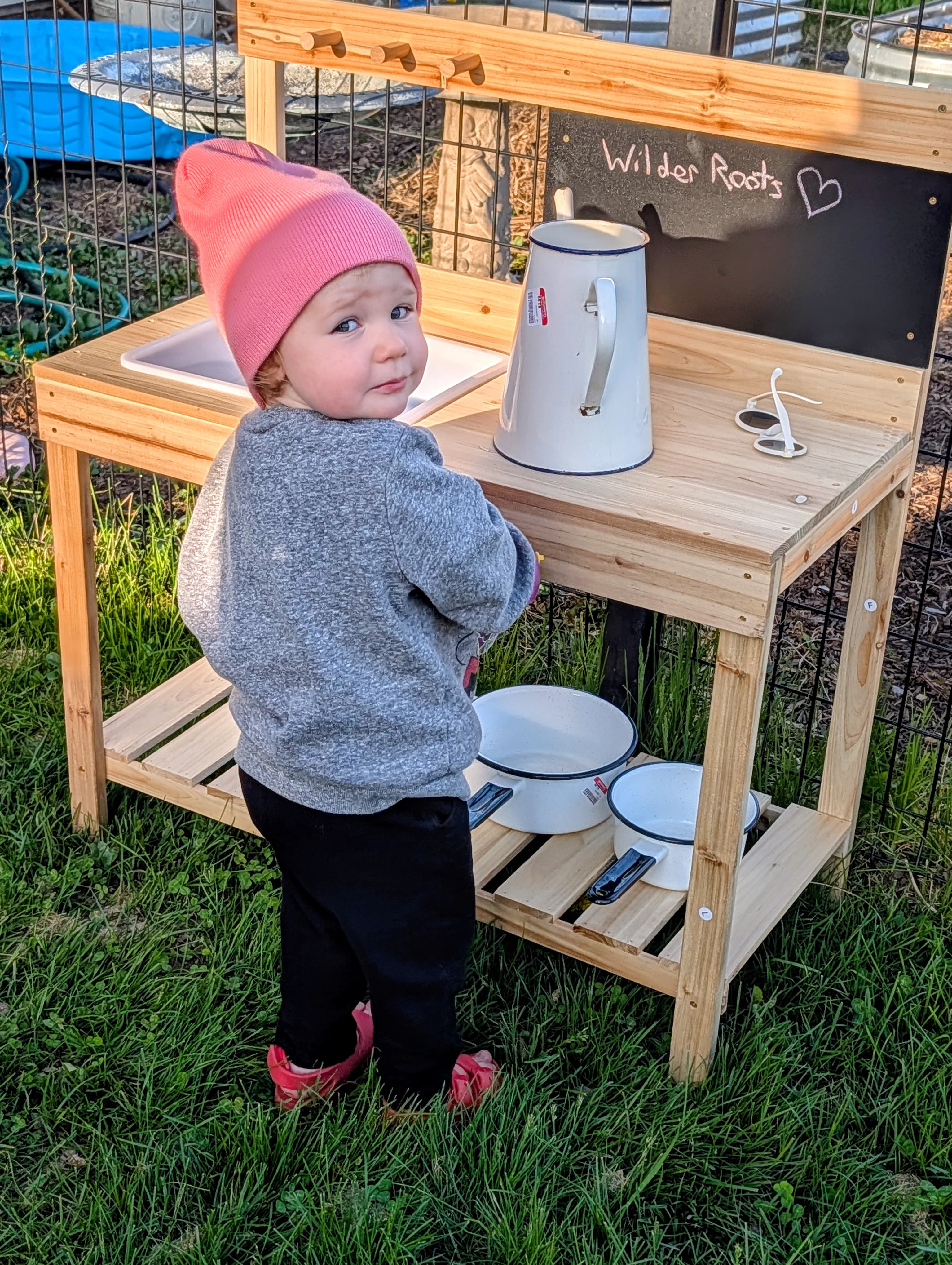 Maeve at her mud kitchen, the chalkboard lettered 'Wilder Roots' — where the brand got its name.